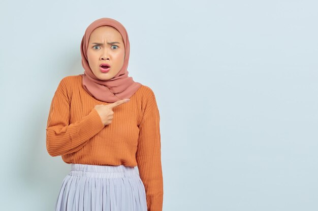 Überrascht schöne asiatische Frau in braunem Pullover und Hijab, die mit dem Finger in den Kopierraum zeigt, der Werbeprodukte zeigt und selbstbewusst in die Kamera blickt, isoliert vor weißem Hintergrund