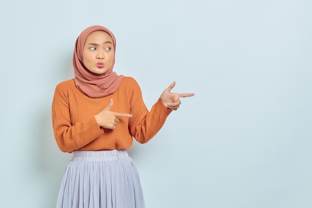 Überrascht schöne asiatische Frau in braunem Pullover und Hijab, die mit dem Finger in den Kopierraum zeigt, der Werbeprodukte zeigt und selbstbewusst in die Kamera blickt, isoliert vor weißem Hintergrund