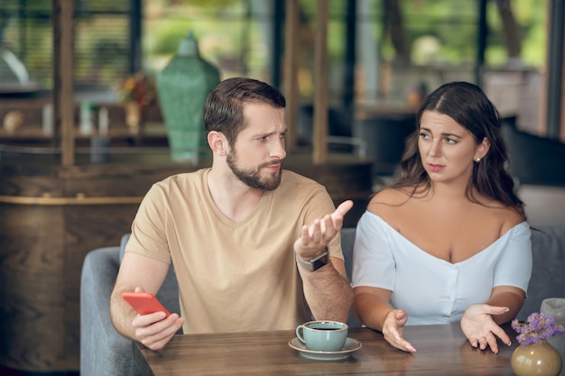 Foto Überraschte beleidigte frau, die fragt und unaufmerksamer mann mit smartphone gestikuliert, das am nachmittag im café sitzt