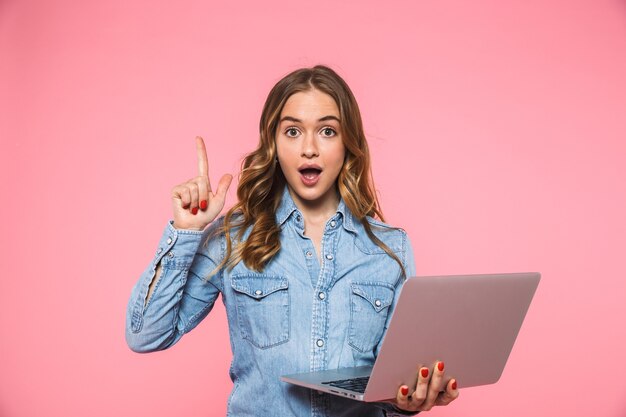 Überraschte blonde Frau, die ein Jeanshemd trägt und eine Idee hat und auf die Vorderseite schaut, während sie einen Laptop über einer rosa Wand hält?