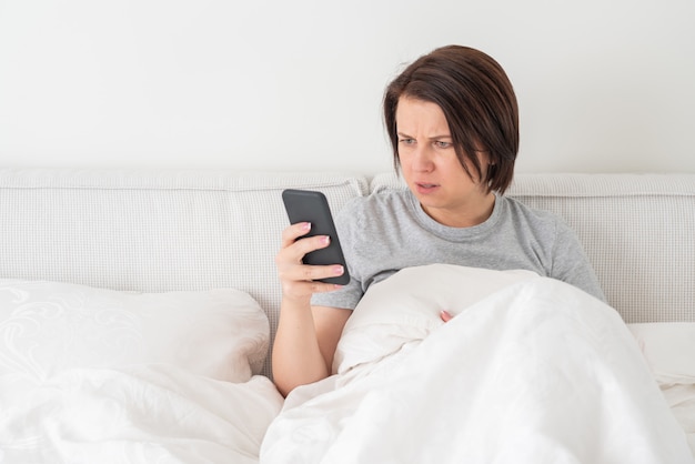 Überraschte frau, die smartphone verwendet, wie sie im bett sitzt, das mit daunendecke bedeckt wird