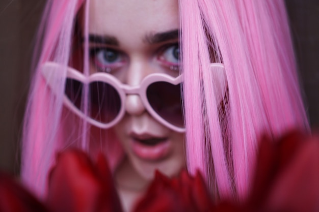 Foto Überraschte frau mit tulpenblumen - rosa haare und herzförmige brille - unscharf, verschwommener hintergrund