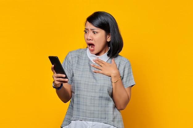 Überraschte junge Asiatin mit Handy mit offenem Mund auf gelbem Hintergrund