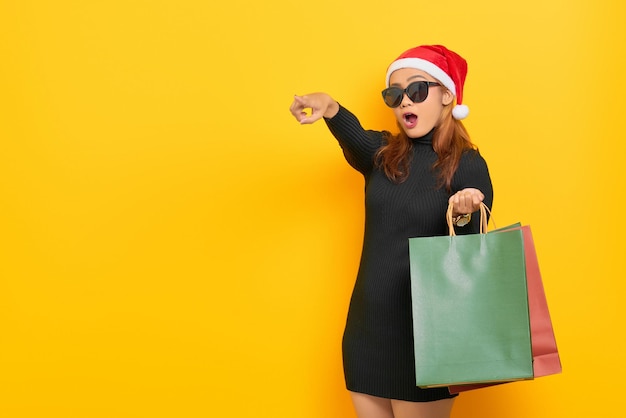 Foto Überraschte junge asiatin mit weihnachtsmann-hut und sonnenbrille mit einkaufstüten, die mit dem finger auf den kopienraum zeigt, der auf gelbem hintergrund isoliert ist