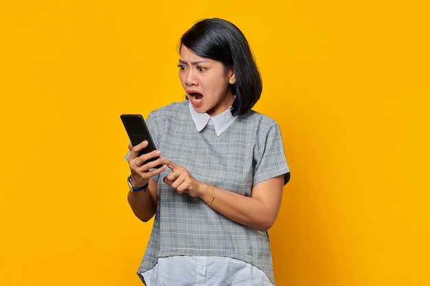 Überraschte junge asiatische Frau, die ein Handy benutzt und Smartphone-Bildschirm auf gelbem Hintergrund schaut