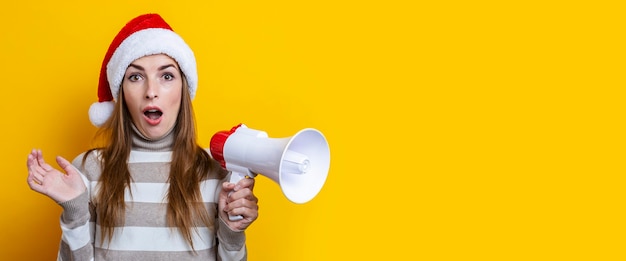 Foto Überraschte junge frau im weihnachtsmann-hut mit einem megaphon auf gelbem grund. banner.