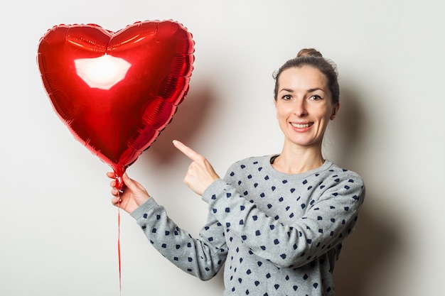 Überraschte junge frau in einem pullover zeigt ihren finger auf den herzluftballon auf einem hellen hintergrund. valentinstag konzept. suche nach einem geliebten menschen.