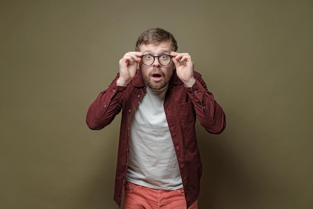 Foto Überraschter bärtiger mann in einem burgunderfarbenen samthemd hält staunend eine brille mit den händen