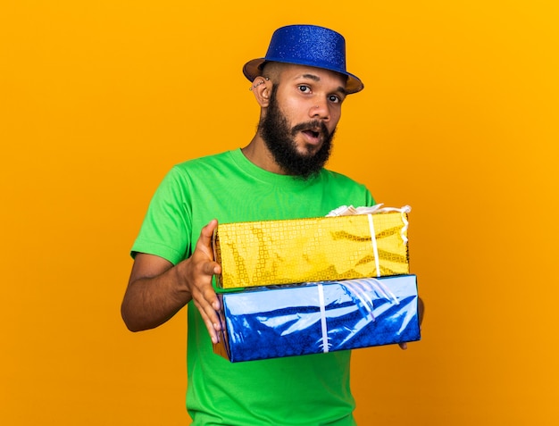 Überraschter junger Afroamerikaner mit Partyhut, der Geschenkboxen isoliert auf oranger Wand hält