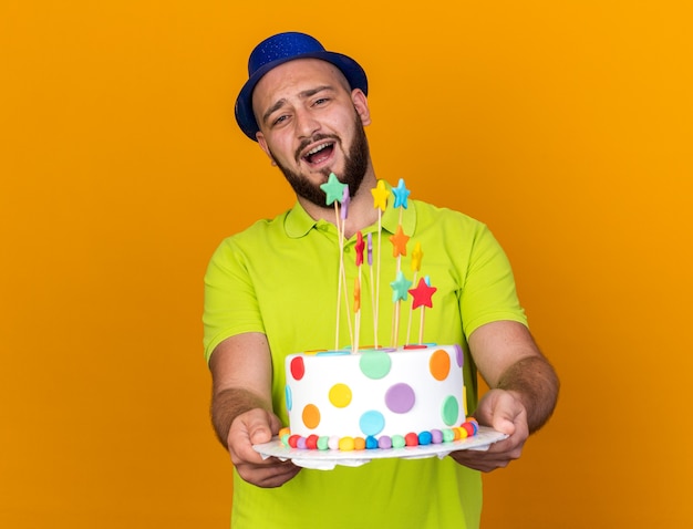 Überraschter junger Mann mit Partyhut, der Kuchen in die Kamera hält