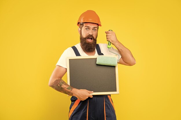 Foto Überraschter mann in helm und arbeitskleidung hält farbe und tafel mit kopierplatz auf gelben hintergrund