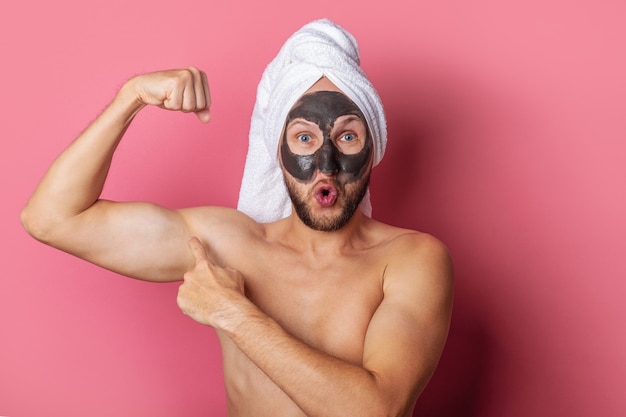 Überraschter nackter Mann mit kosmetischer Maske auf seinem Gesicht, zeigt Finger, der Muskel auf rosa Hintergrund zeigt.