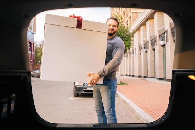 UFA, RUSIA - 13 DE SEPTIEMBRE DE 2020. Retrato de un repartidor sonriente sosteniendo una gran caja festiva con lazo rojo cerca del coche al aire libre en la calle de la ciudad y mirando la cámara. Mensajero masculino entregando un regalo al cliente.