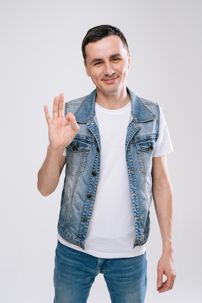 UFA, RUSIA - 31 DE MARZO DE 2020. Joven hombre guapo sonriente con ropa de denim posando mostrando un gesto OK con las manos sobre fondo de pared blanca, retrato de estudio. Concepto de apostar emociones brillantes