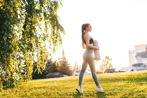 UFA RUSSLAND 7. SEPTEMBER 2020 Aktive junge Frau, die Yogamatte hält und im Stadtpark lächelt, bereit, Yoga mit einem Glücksgefühl im Hintergrund von hellem Sonnenlicht zu praktizieren Konzept eines gesunden Lebensstils