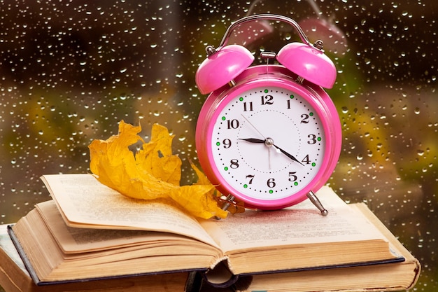 Uhr und trockenes Ahornblatt auf einem Buch vor dem Hintergrund des Fensters bei Regenwetter im Herbst des Abends.