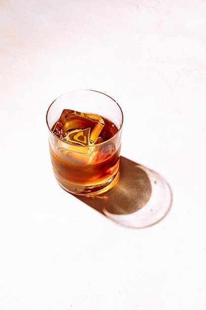 Uísque ou bourbon em um copo de rochas com um grande cubo de gelo, tiro com luz dura e sombras duras, espaço de cópia de fundo brilhante