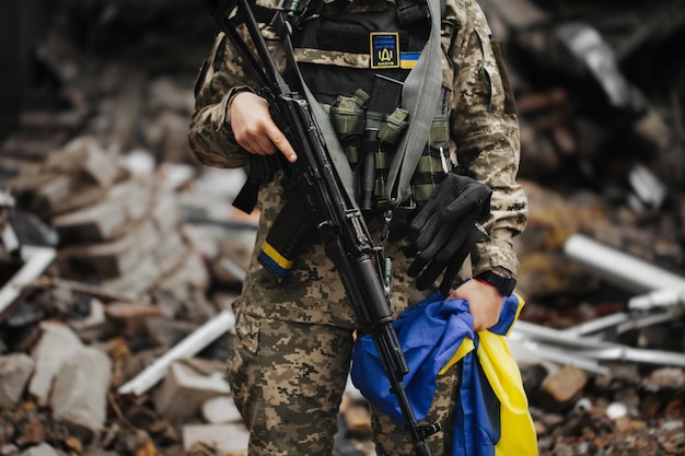 Foto ukrainische militärfrau mit der ukrainischen flagge in ihren händen auf dem hintergrund eines explodierten hauses