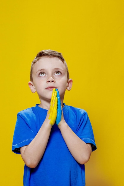 Ukrainischer Junge betet für ukrainische Kinder gegen den Krieg Ein Junge in einem blauen T-Shirt auf gelbem Hintergrund faltete seine Hände, die in der ukrainischen Flagge zum Gebet gemalt waren