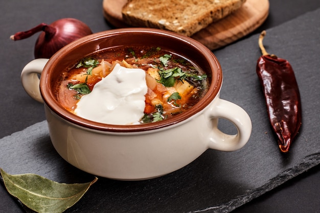 Ukrainischer traditioneller Borschtsch in Porzellanschüssel mit Sauerrahm, Roggenbrot, Zwiebeln und Chilipfeffer auf Steinbrett. Ansicht von oben.