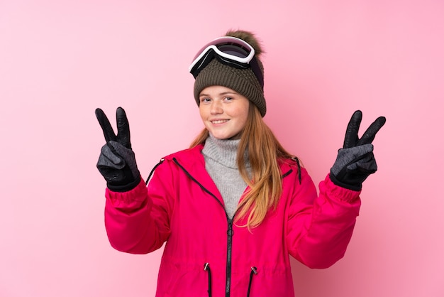 Ukrainisches Jugendlichskifahrermädchen mit Snowboardinggläsern über lokalisiertem Rosa lächelnd und Siegeszeichen zeigend