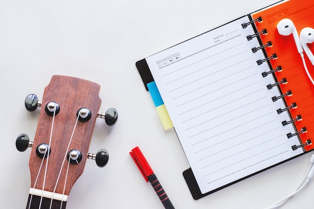 Foto ukulele com caderno espiral aberta, caneta e fones de ouvido no fundo branco