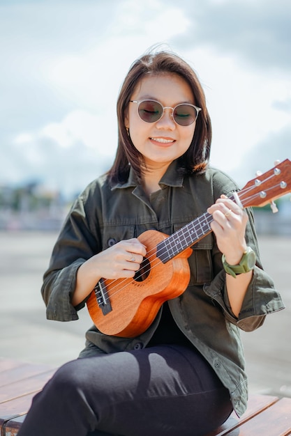 Ukulele der jungen schönen asiatischen Frau mit Jacke und schwarzer Jeans spielen, die im Freien posiert