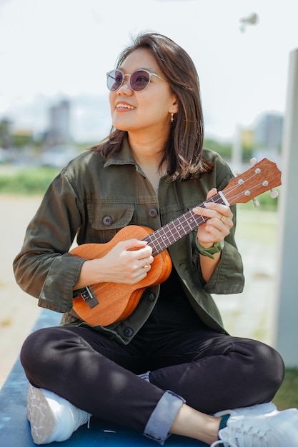 Ukulele der jungen schönen asiatischen Frau mit Jacke und schwarzer Jeans spielen, die im Freien posiert