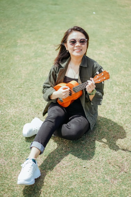 Ukulele der jungen schönen asiatischen Frau mit Jacke und schwarzer Jeans spielen, die im Freien posiert