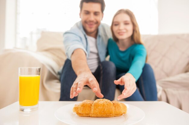 El último croissant. Hermosa joven pareja sentada en el sofá y estirando las manos hasta el último croissant en el plato