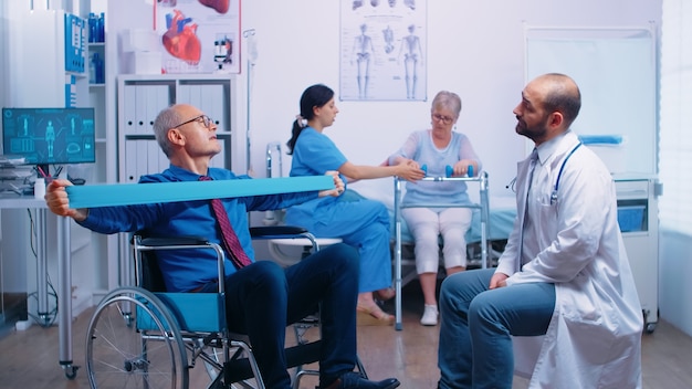 Último homem em cadeira de rodas, exercitando a força muscular com elástico, sob supervisão médica, em uma clínica ou hospital de recuperação moderna programa de fisioterapia inválido, reabilitação de lesões de saúde