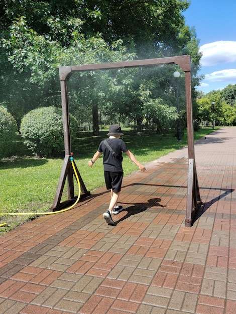 Um adolescente caminha sob um quadro de resfriamento com salpicos de água em um parque da cidade Vista traseira