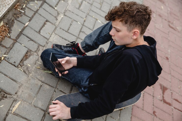 Um adolescente de cabelos cacheados, de aparência europeia, usando um capuz preto, sentado em um beco de skate e ...
