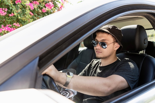 Um adolescente dirige um jovem moderno com óculos escuros da moda e barba, dirige um carro branco ...