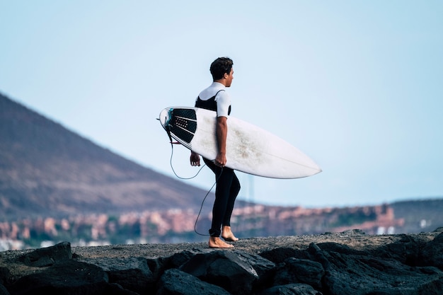 Foto um adolescente entrando na água com sua prancha debaixo do braço pronto para surfar - atleta ativo vai treinar na água - homem com roupa de neoprene