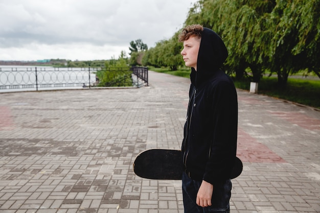 Um adolescente europeu de cabelos cacheados em um moletom preto com um skate nas mãos está de pé na margem ...