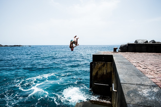 Um adolescente jovem e feliz pulando de um penhasco para a água do mar ou oceano em um dia quente de verão, desfrutando da praia. estilo de vida ativo e bonito ao ar livre