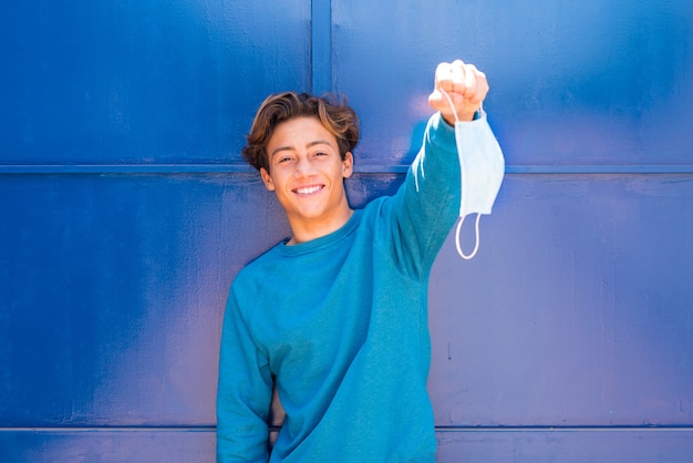 Um adolescente segurando uma máscara médica e cirúrgica após quarentena e bloqueio em casa sorrindo e feliz por vencer o covid-19 ou coronavírus - sweather azul e parede azul ao fundo
