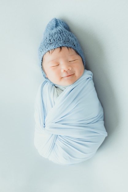 Um adorável adorável sorridente asiático bebê recém-nascido com chapéu de malha azul e wrapprd em casulo, dormindo na cama
