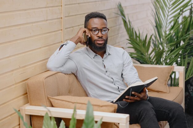 um afro-americano bonito e jovem em um escritório ou coworking falando ao telefone e tomando notas em um notebook trabalhando em um laptop. empresário masculino afro-americano, comerciante ou financista, moderador