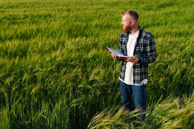 Um agricultor barbudo com um notebook fica no centro de um grande campo de trigo Preparação da colheita durante a crise alimentar no mundo