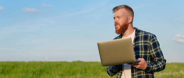 Um agricultor barbudo e brutal com um laptop está de pé em um campo com trigo Inspeção da futura colheita Fotografia panorâmica