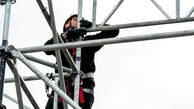 Um alpinista industrial usando um capacete e um cinto de proteção é amarrado com uma corda a um suporte em alta altitude Um especialista monta uma estrutura de palco a partir de um sistema modular de andaimes metálicos