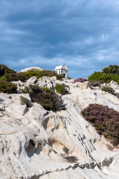 Um antigo e pequeno santuário localizado nas rochas perto da costa do mar Egeu, arbustos ao redor, céu nublado, Grécia