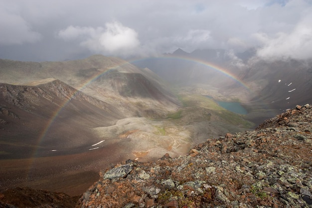 Um arco-íris completo em um vale fotografado de cima de uma passagem na montanha. Nuvens sombrias no céu. Abaixo você pode ver o lago. Horizontal.