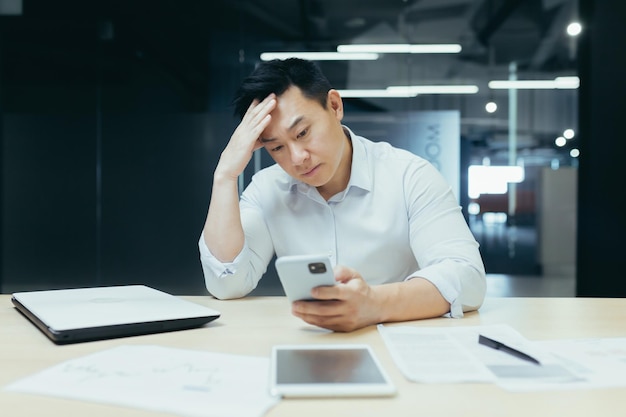 Um asiático confuso segurando um smartphone se sente preocupado pensando na mensagem recebida no celular