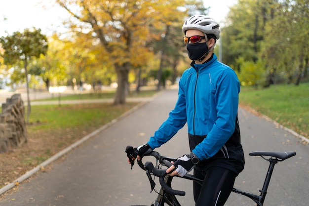 Um atleta de óculos e uma máscara protetora fica no parque em uma estrada de bicicleta com uma bicicleta. Esportes em quarentena e recreação ao ar livre.