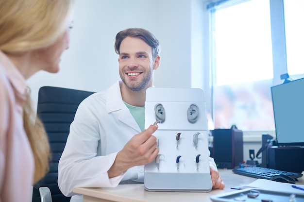 Um audiologista bonito e sorridente mostrando uma diversidade de aparelhos auditivos para uma mulher loira