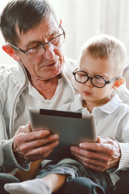 Um avô idoso e seu neto estão usando um tablet juntos, sorrindo e se divertindo ou assistindo a um vídeo engraçado na Internet, sentados em uma cadeira