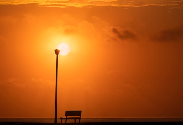Um banco de madeira e uma lâmpada de rua com uma vista de fundo de um pôr do sol vermelho no pôr do sol e um sol brilhante.